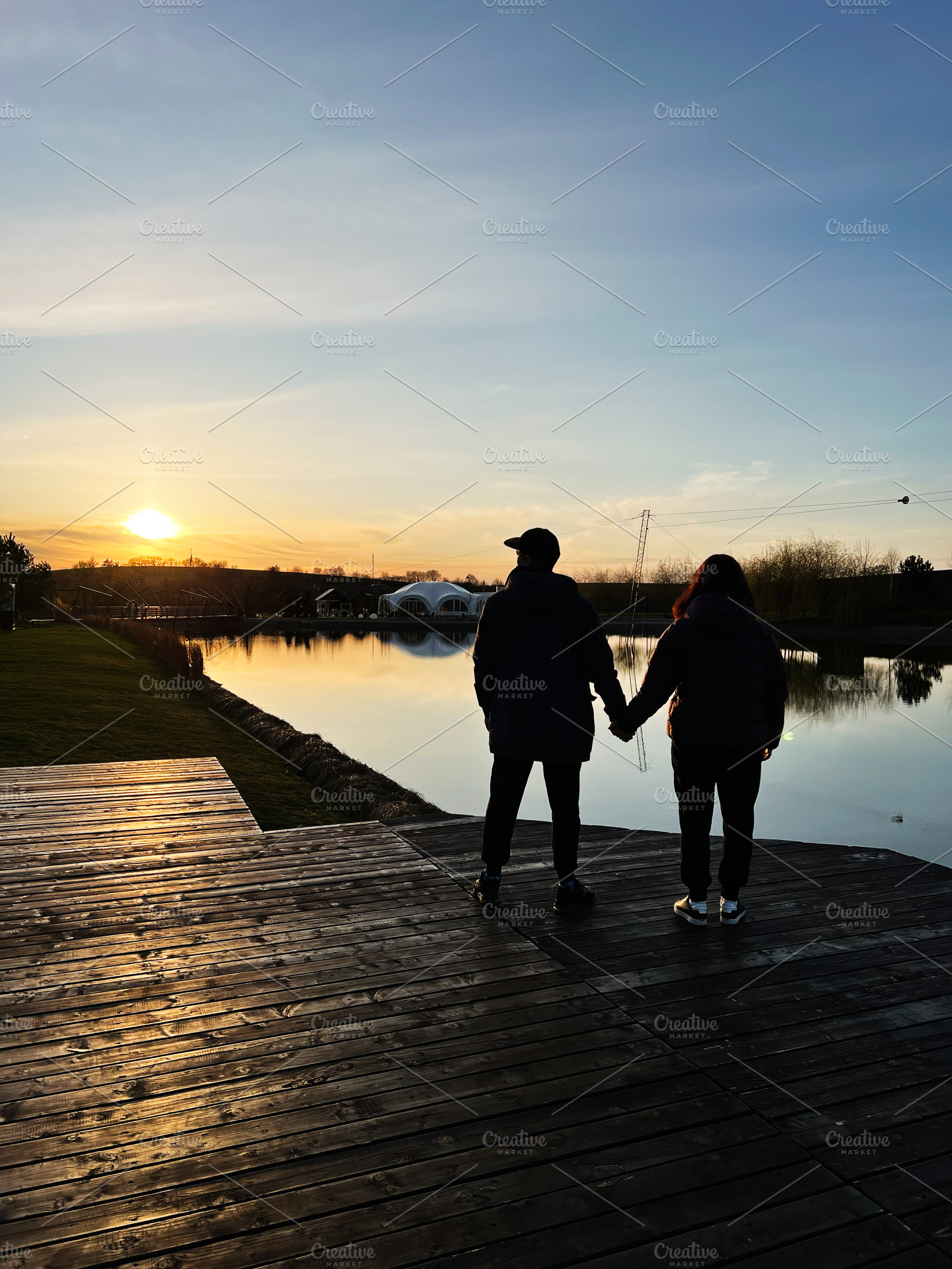 Couple enjoying a serene lakeside sunset while holding hands on a wooden deck, a Photo by AS photostudio