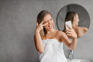 Playful woman taking selfie on smartphone while enjoying beauty morning routine, a Photo by Prostock-Studio