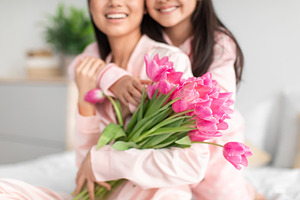 Glad teenage girl hugging millennial japanese lady in pajama giving bouquet of, a Photo by Prostock-Studio
