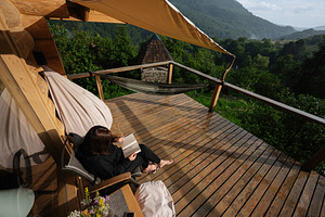 Woman reading a book on a wooden balcony overlooking a lush forest, a Photo by AS photostudio