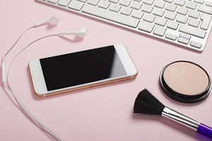 Smartphone headphones and computer keyboard next to makeup on pink and blue