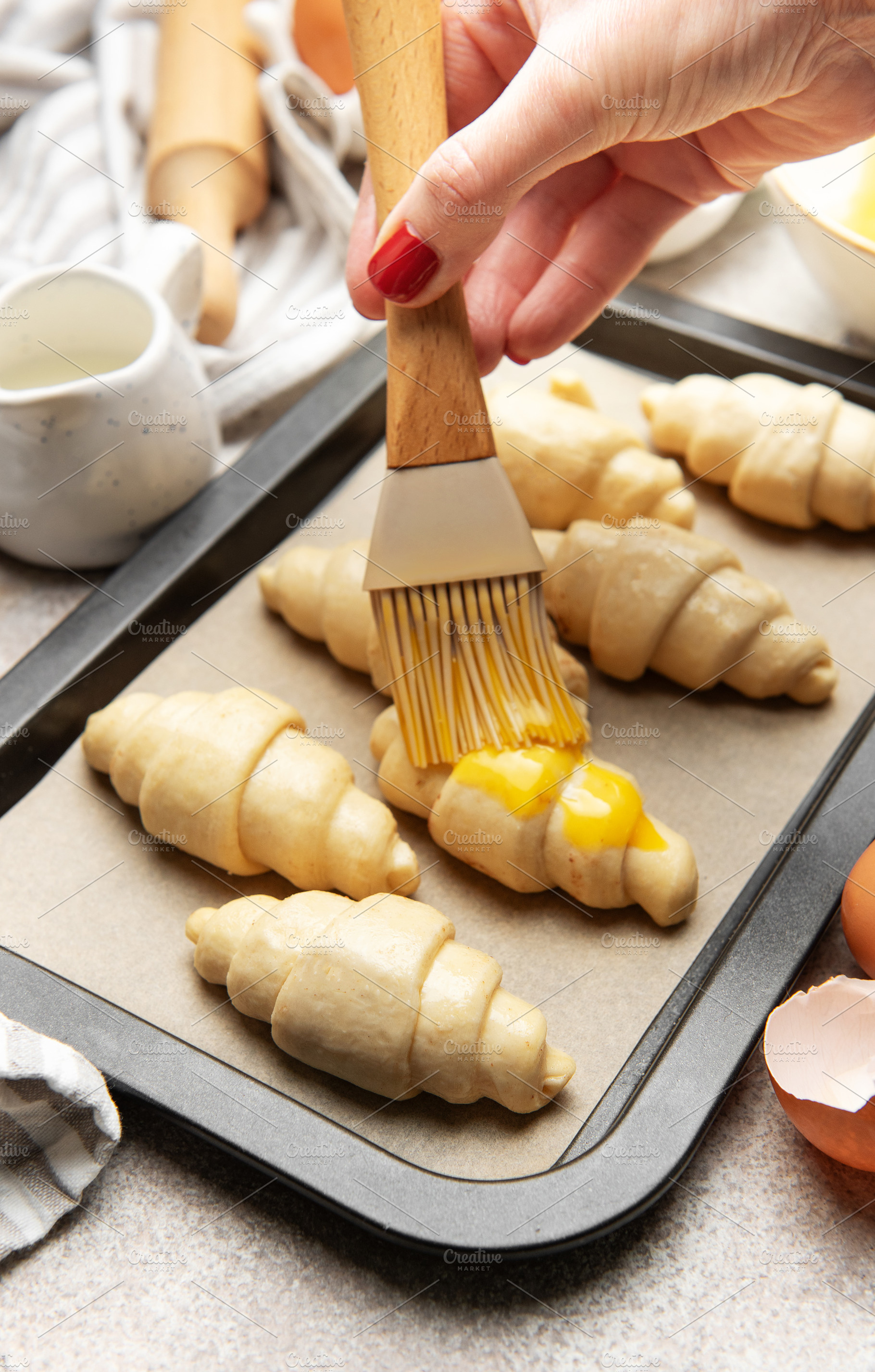 Baker brushing egg yolk on raw croissants on baking sheet, a Food & Drink Photo by Olena Rudo