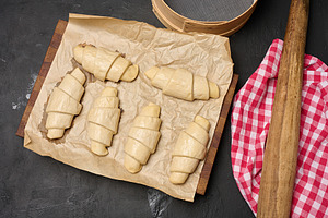 Uncooked croissant blanks on parchment paper and kitchen utensil, a Food & Drink Photo by ndanko