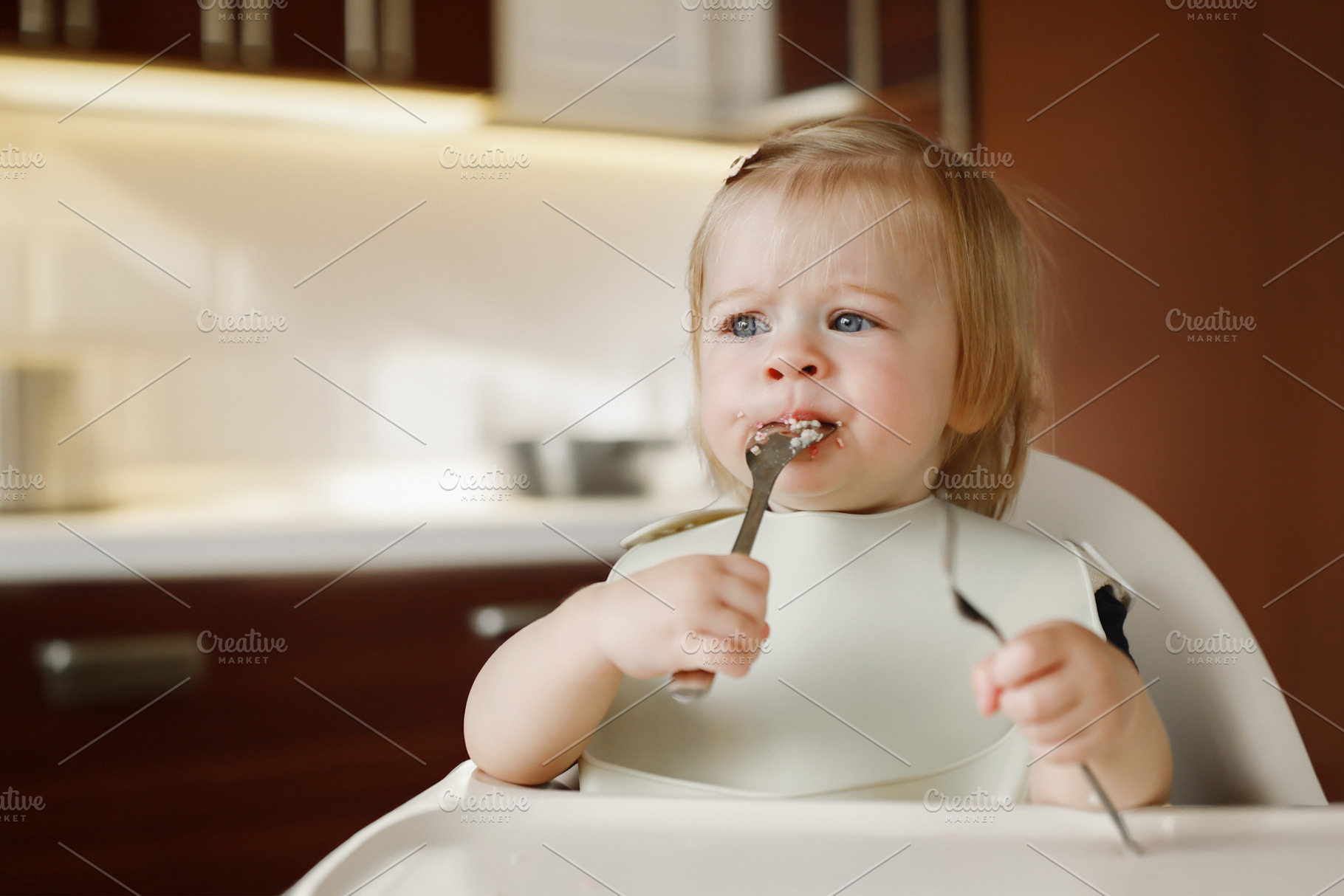 Infant girl or toddler sitting on high chair. Eating food by herself using spoo, a Person Photo by Lenor4ik