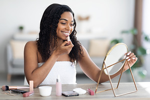 Attractive curly black woman applying lipstick, home interior