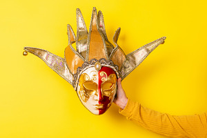 Hand holding ornate Venetian jester mask against vibrant yellow, a Holiday Photo by Ales Munt