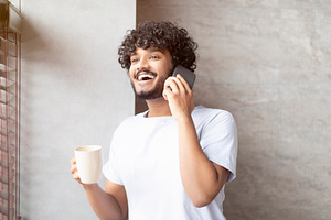Happy indian man having phone conversation, standing in bathroom with cup in ha, a Person Photo by Prostock-Studio