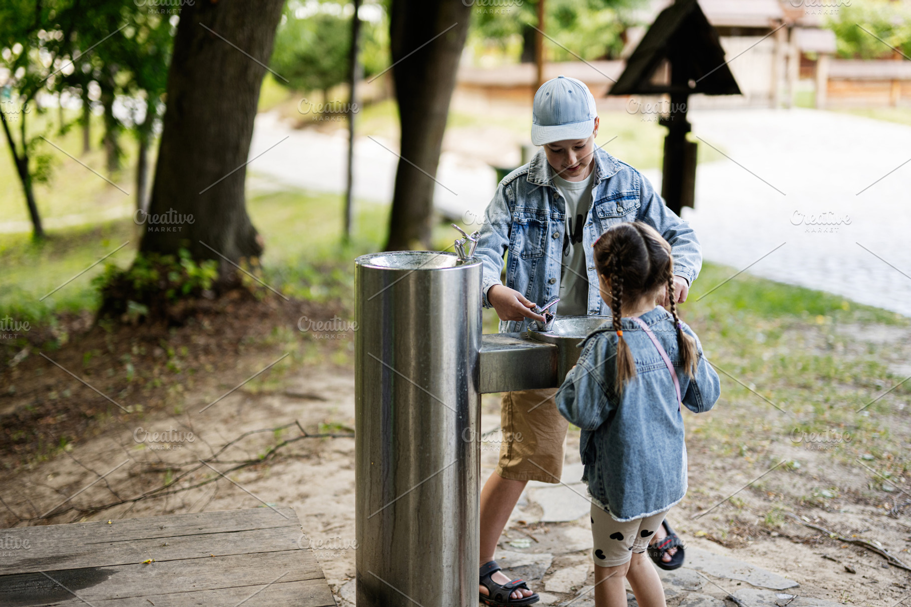 Two children using an outdoor water fountain in a natural park setting, a Person Photo by AS photostudio