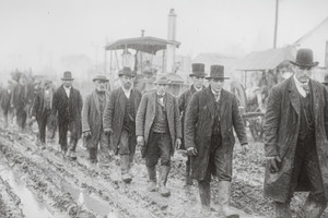 Men in top hats and work coats walking through a muddy road in a vintage farmin, a Photo by AS photostudio
