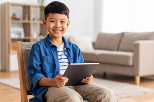 Boy with a tablet sitting on chair at living room while smiling, a Person Photo by DanFLCreativo