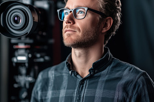 A thoughtful motion designer with glasses stands in front of a camera, wearing, a Person Photo by Textures & Backgrounds