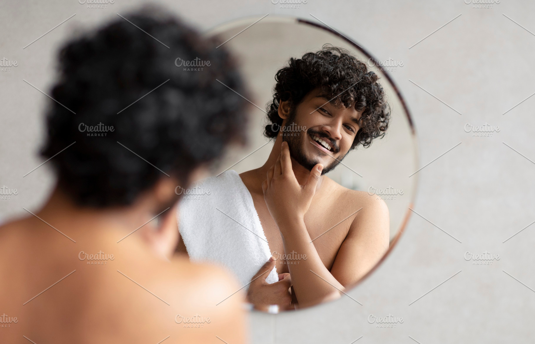 Young indian man looking in mirror in bathroom and touching beard, handsome guy, a Person Photo by Prostock-Studio
