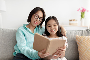 Loving Asian grandmother reading book with her cute little granddaughter on cou, a Photo by Prostock-Studio