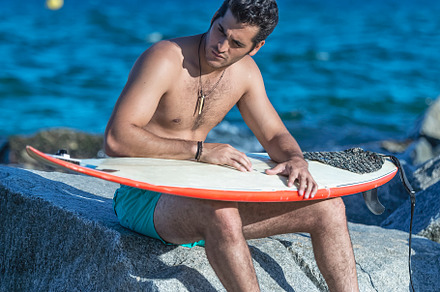 Serious man sitting on rocks with surfboard.