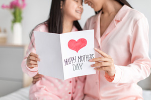 Glad happy teen chinese girl and millennial female in pajamas hold postcard in, a Photo by Prostock-Studio