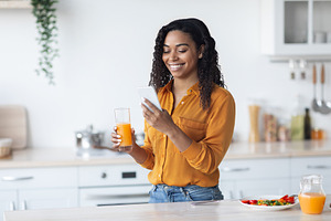 Positive pretty black lady holding glass of juice and smartphone