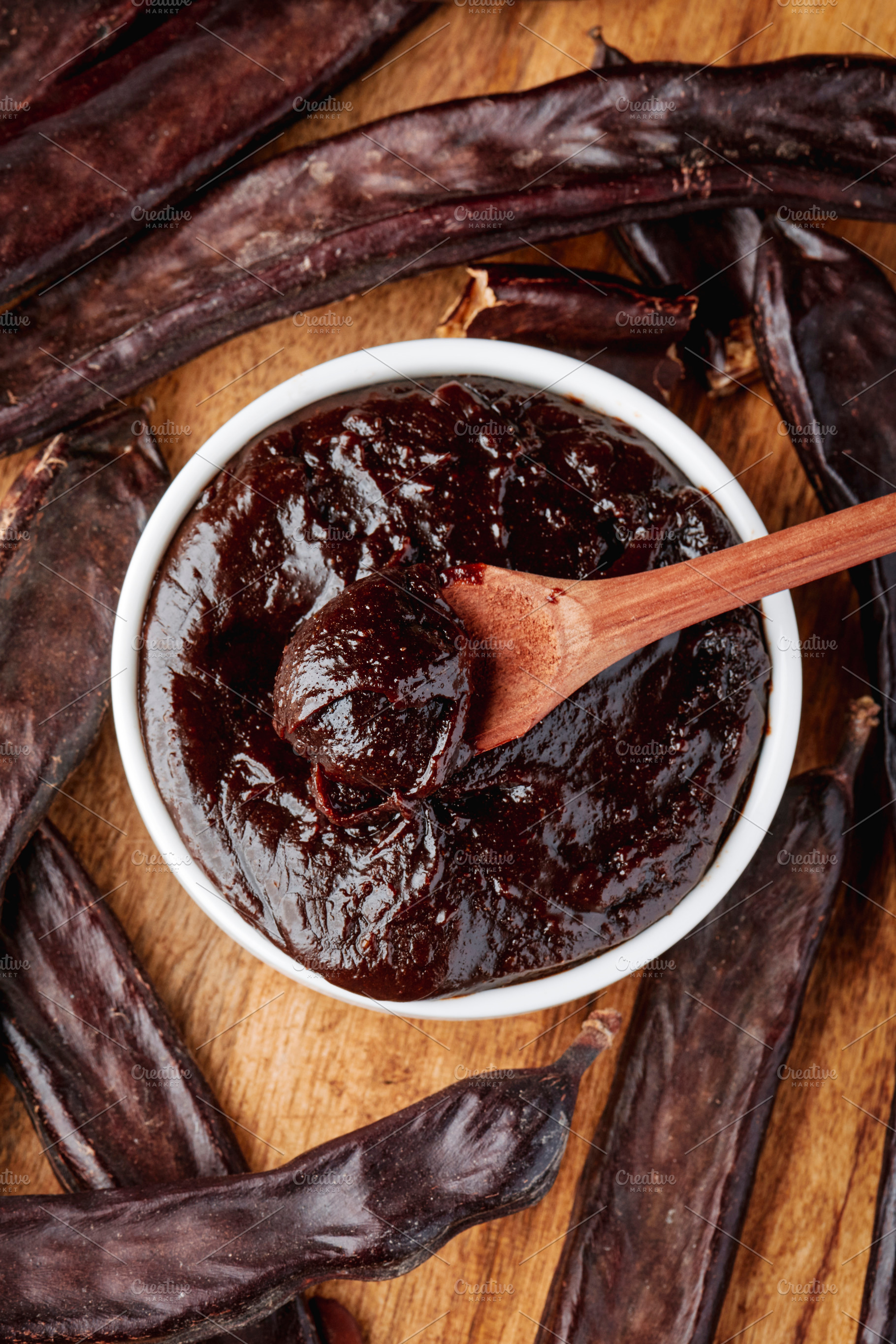 bowl of carob spread with spoon surrounded by pods, a Photo by Nito