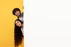 Excited indian couple standing behind white advertisement board and peeking out, a Photo by Prostock-Studio