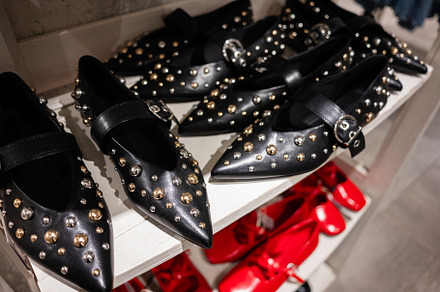 Black leather flats with studs displayed on a shelf with red shoes nearby, a Photo by AS photostudio