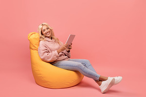 Happy lady using digital tablet while sitting in beanbag chair over pink backgr, a Photo by Prostock-Studio