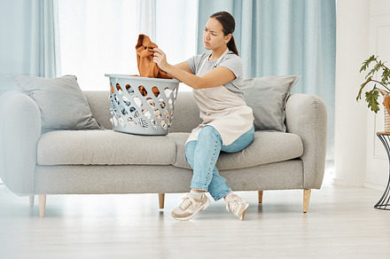 Asian woman, laundry or clothes basket on sofa in house living room and looking confused at dirty stains. Stress, anxiety or worry of person with washing clothing or cleaning fabric in home interior, a Photo by Peopleimages.com