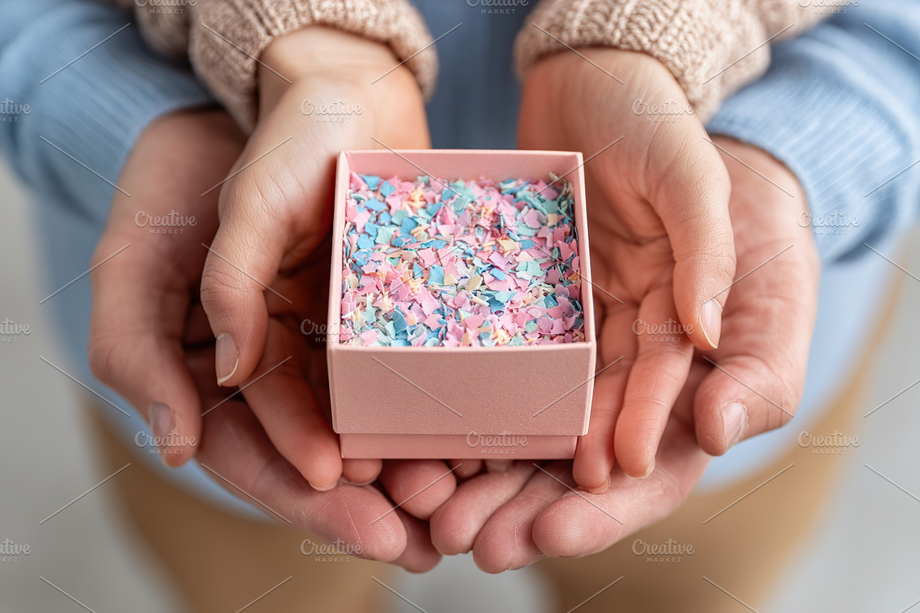 Parents' hands gently holding a small pink box filled with colorful confetti, a Photo by AlyaAnd