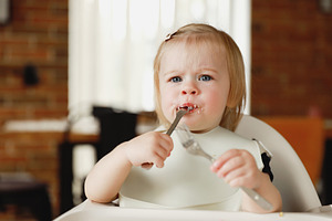 Infant girl or toddler sitting on high chair. Eating food by herself using spoo, a Person Photo by Lenor4ik