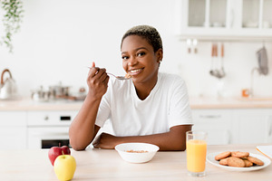 Smiling young attractive black woman eating porridge sits at table with cookies, a Person Photo by Prostock-Studio