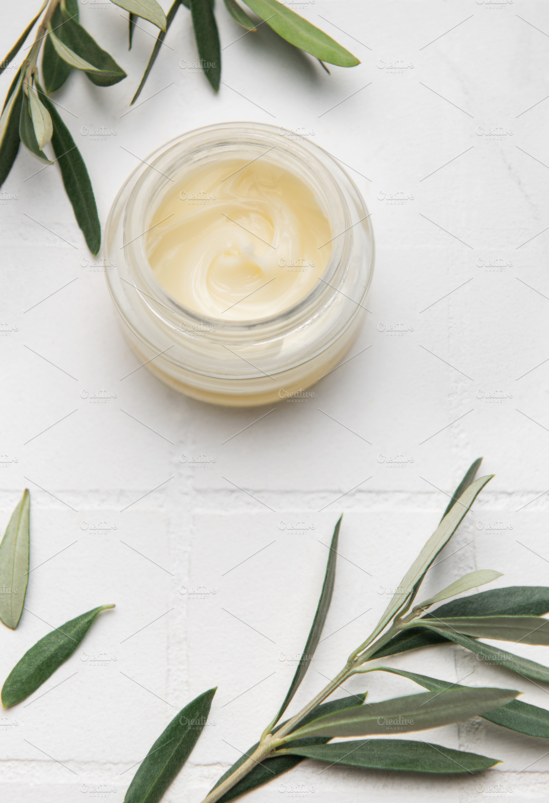 Moisturizing cream surrounded by olive branches on white surface, a Health & Medical Photo by Olena Rudo