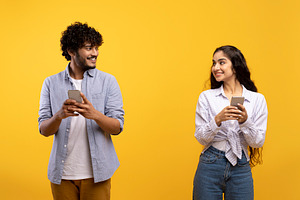 Cheerful young indian man and woman in casual outfits holding smartphones, look, a Photo by Prostock-Studio
