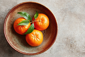 Rustic ceramic bowl filled with fresh mandarins and green leaves on a textured, a Food & Drink Photo by AlyaAnd