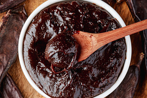 bowl of carob spread with spoon surrounded by pods, a Photo by Nito