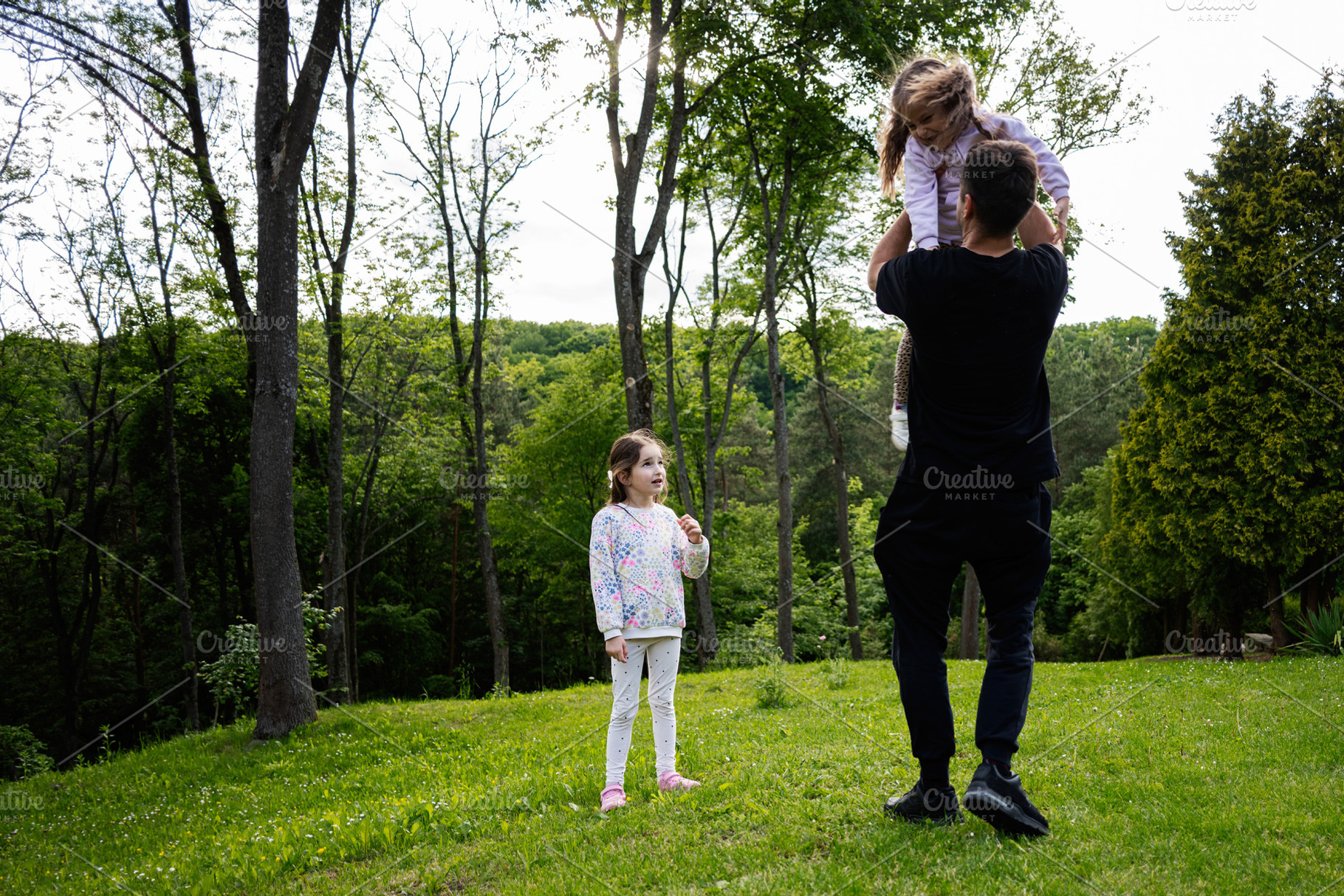 Father lifting daughter in park while another child watches, surrounded by tree, a Person Photo by AS photostudio