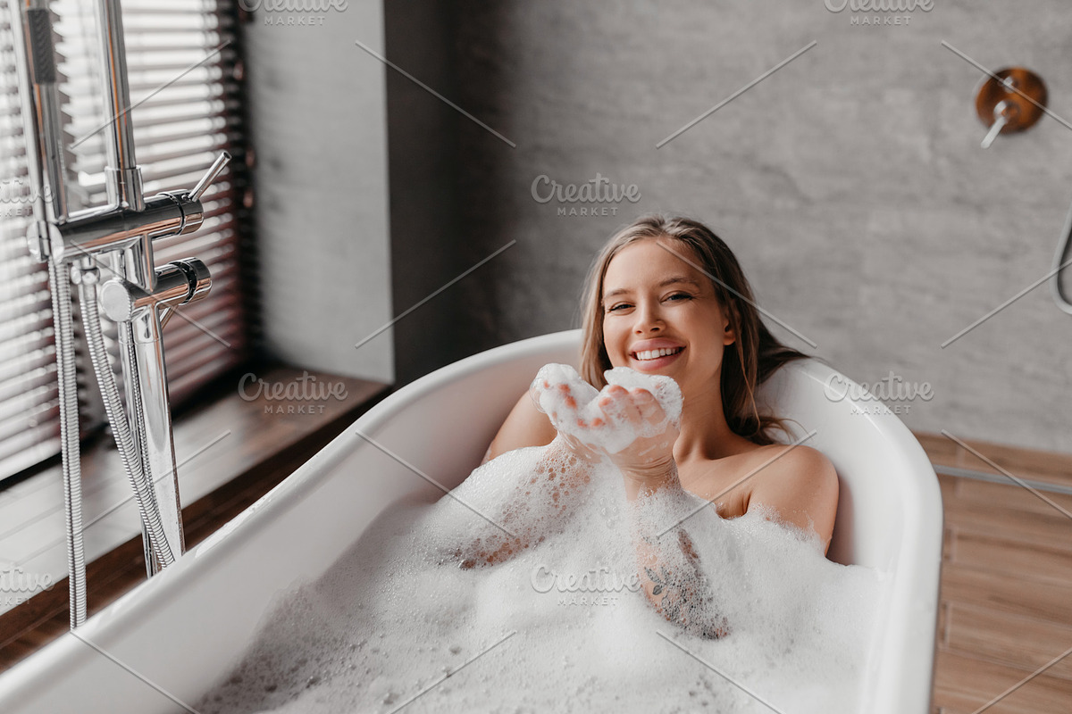 Young pretty woman relaxing in bathtub, playing and blowing foam, looking and s, a Photo by Prostock-Studio (Photo 2 of 2)