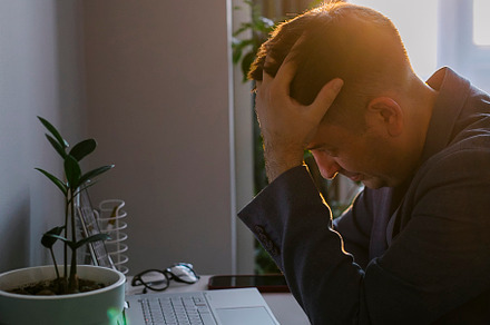 Young man sitting at office table and looking at laptop screen, touch forehead, a Business Photo by Anikona