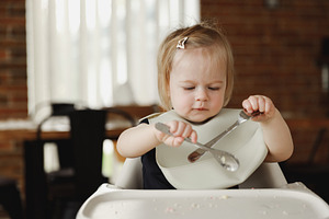 Infant girl or toddler sitting on high chair. Eating food by herself using spoo, a Person Photo by Lenor4ik