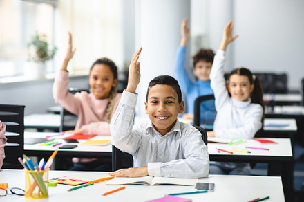 Diverse small schoolkids raising hands at classroom, a Photo by Prostock-Studio