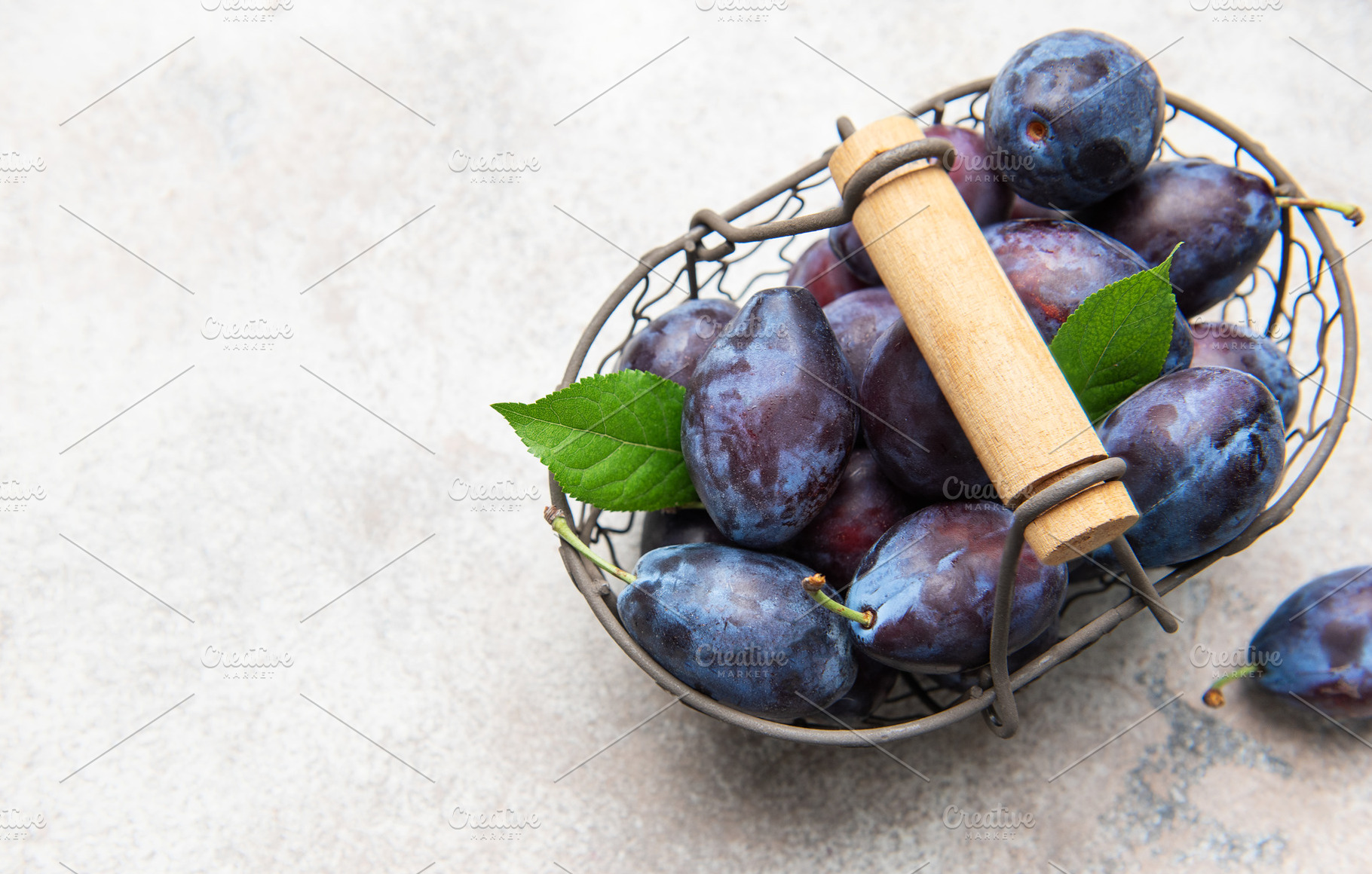 Fresh purple plums in wire basket on concrete, a Food & Drink Photo by Olena Rudo