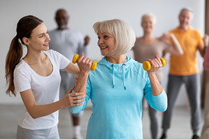 Female fitness coach helping senior woman while exercising, a Photo by Prostock-Studio
