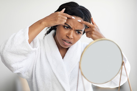 Young black lady standing in front of mirror, removing blackheads, a Photo by Prostock-Studio