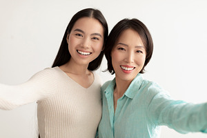 Happy young Asian woman and her mature mother taking selfie together on white s, a Photo by Prostock-Studio