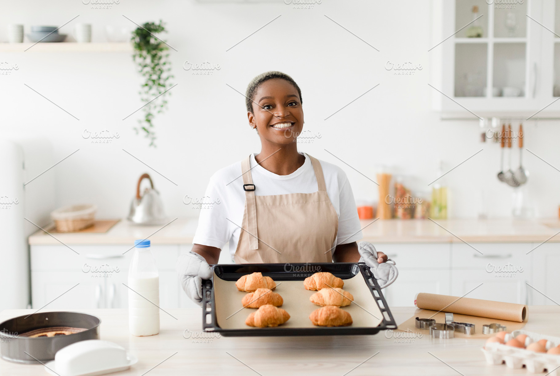 Satisfied young cute black woman in apron shows ready croissants in modern kitc, a Person Photo by Prostock-Studio