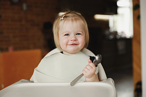 Infant girl or toddler sitting on high chair. Eating food by herself using spoo, a Person Photo by Lenor4ik