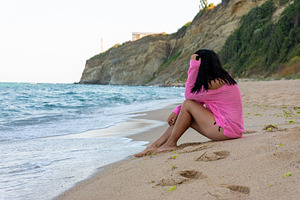 Woman in pink sweater sitting on a sandy beach near ocean waves, with cliffs an, a Holiday Photo by Ales Munt