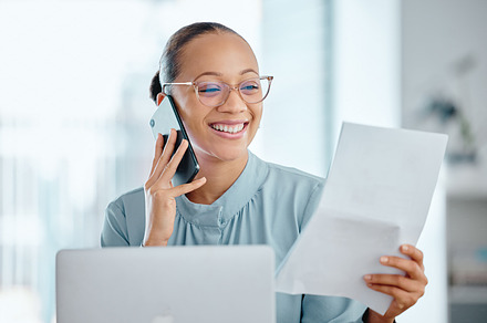 Happy business woman talking on a phone call, discussing contract with a client or colleague. Female executive holding and reading documents or paperwork, sitting in front of a laptop at office desk., a Photo by Peopleimages.com