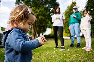 Young child holding an object outdoors with family in blurred background, a Photo by AS photostudio