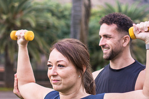 Male personal trainer assisting woman with dumbbell exercise outdoors