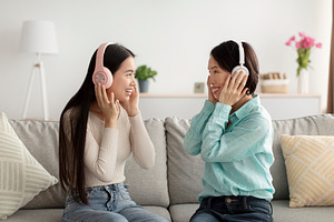 Happy young Asian woman and her mature mother in headphones listening to music, a Photo by Prostock-Studio