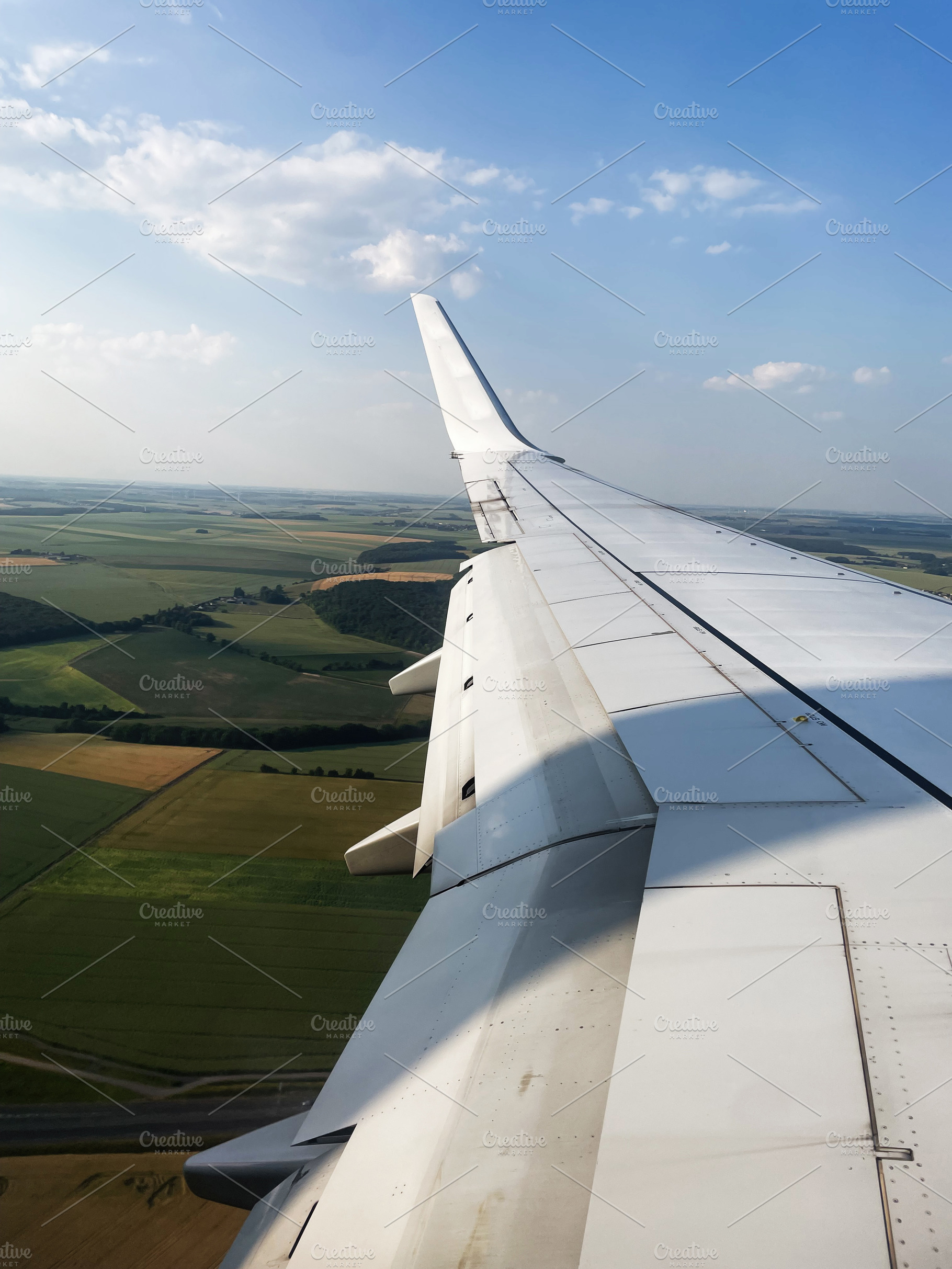 Airplane wing mid-flight above green fields under a blue sky, a Photo by AS photostudio