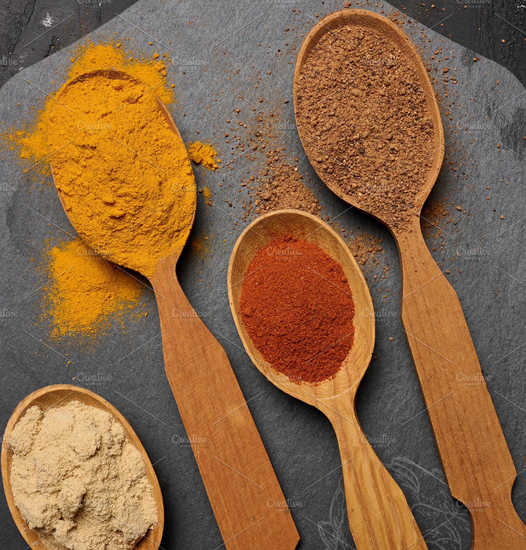 Multicolored ground spices in wooden spoons on a black board,, a Food & Drink Photo by ndanko